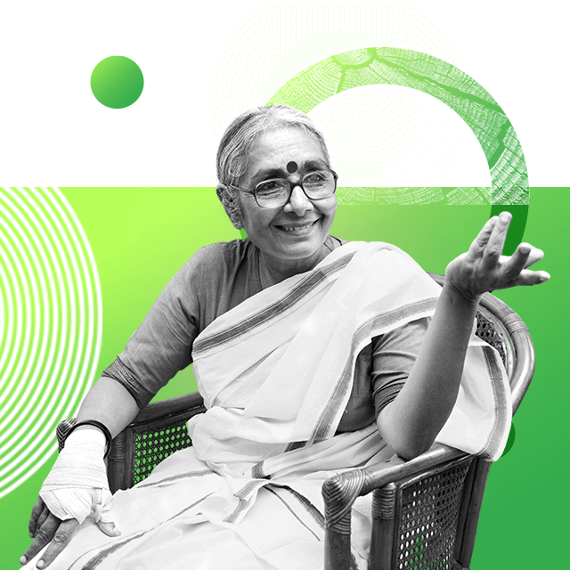 Social activist Aruna Roy is wearing a saree with her hair tied back behind her shoulders. She sits on a rattan chair with her right hand bandaged and her left hand lifted in the air as if she is in mid conversation.