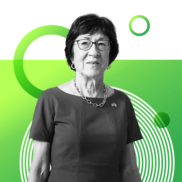 Senator Susan Collins with short hair and glasses, wearing a dark tailored short-sleeved dress with a chunky metal necklace and a pin with two small flags on her lapel.