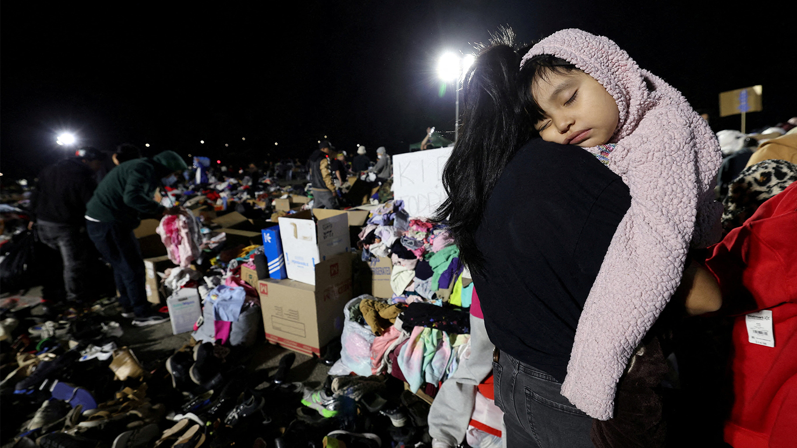 A small girl lies asleep on her mother's shoulder in front of piles of donations at a distribution center for people escaping the Eaton fire.