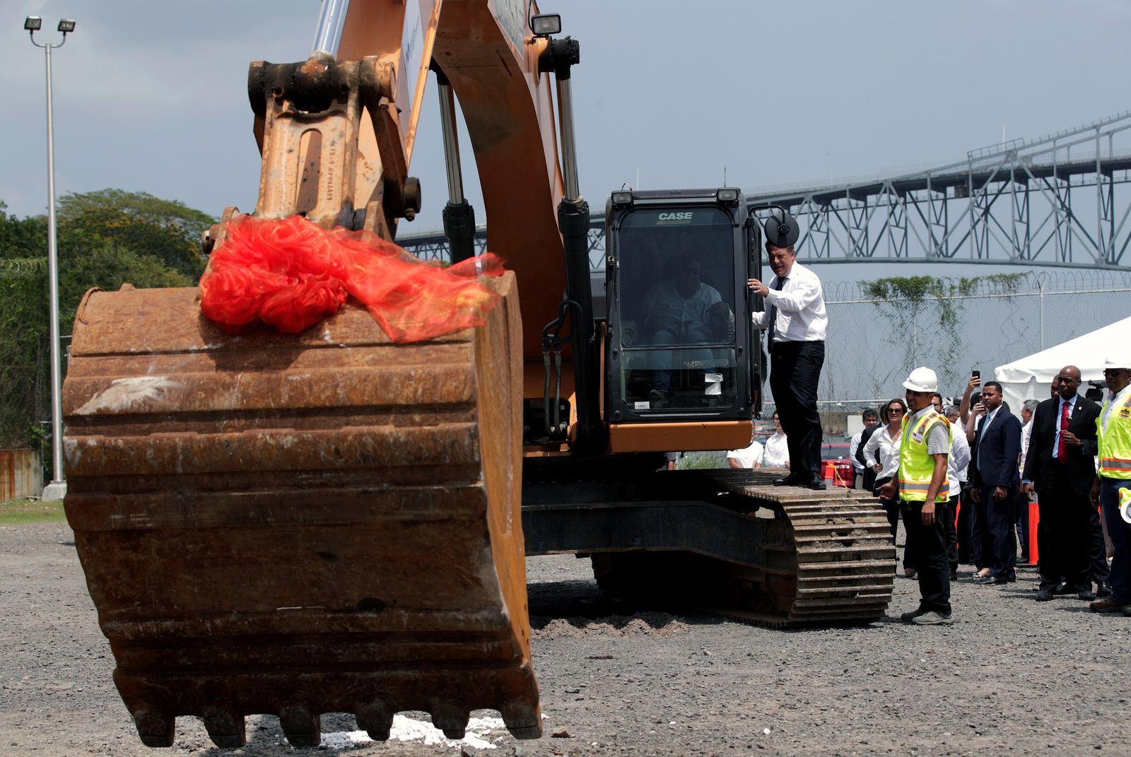 A crowd in either suits or hi-vis vests watches as a digger begins work on the fourth bridge over the Panama Canal with another bridge visible behind. A man in a shirt and tie is standing on the digger track talking to the driver and some orange ribbon can be seen hanging from the digger's scoop after it cut through.