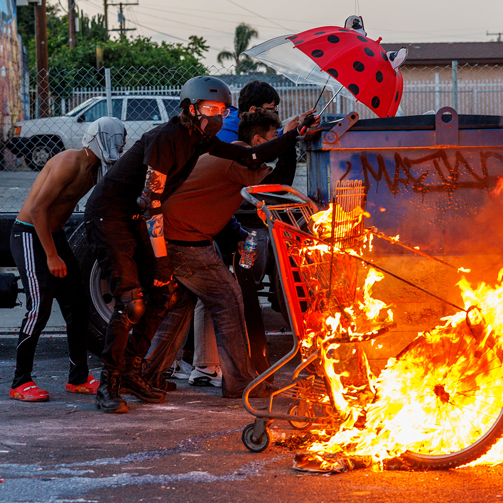 Protesters hide behind a dumpster near a set fire as LA County Sheriff deputies shoot projectiles to keep demonstrators from advancing after ICE raids at a nearby Home Depot and the Garment District