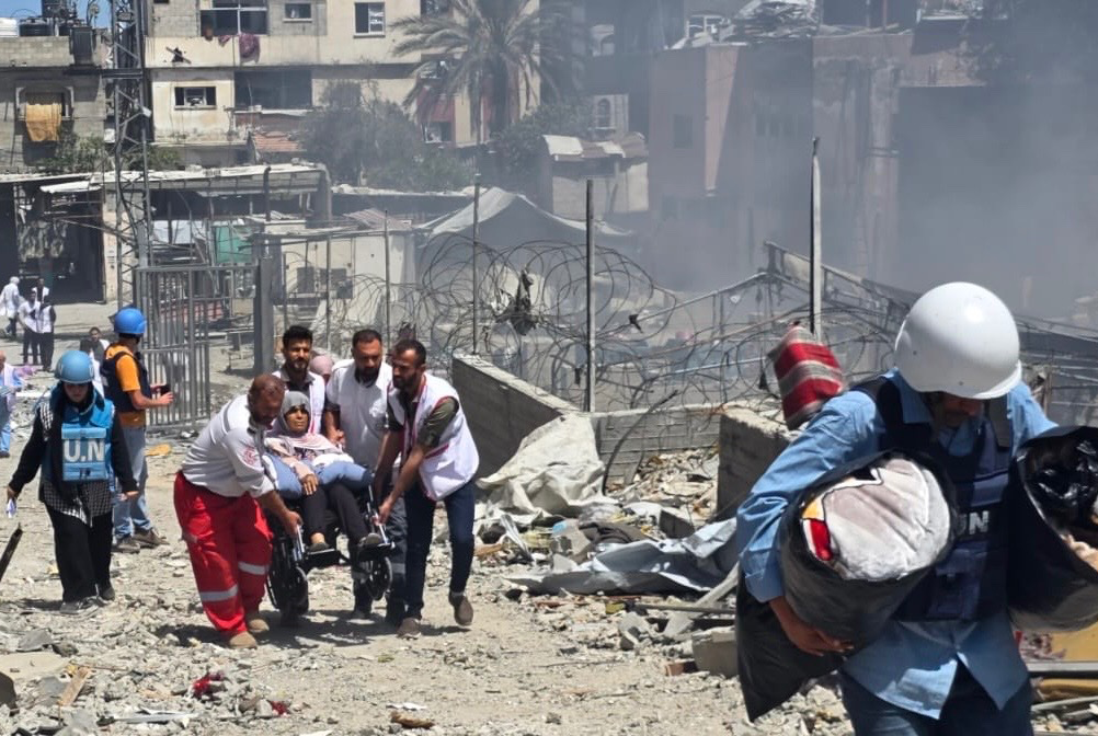 Medical workers and UN staff assist an injured person on a stretcher through rubble and debris in a heavily damaged urban area of Gaza. Smoke rises in the background, and barbed wire lines part of the street.
