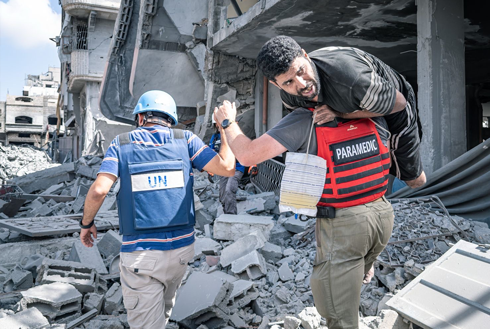 A paramedic wearing a red vest carries a man over his shoulder through the rubble of a destroyed building in Gaza. A UN worker in a blue vest and helmet walks ahead, guiding the way through the debris-strewn street.