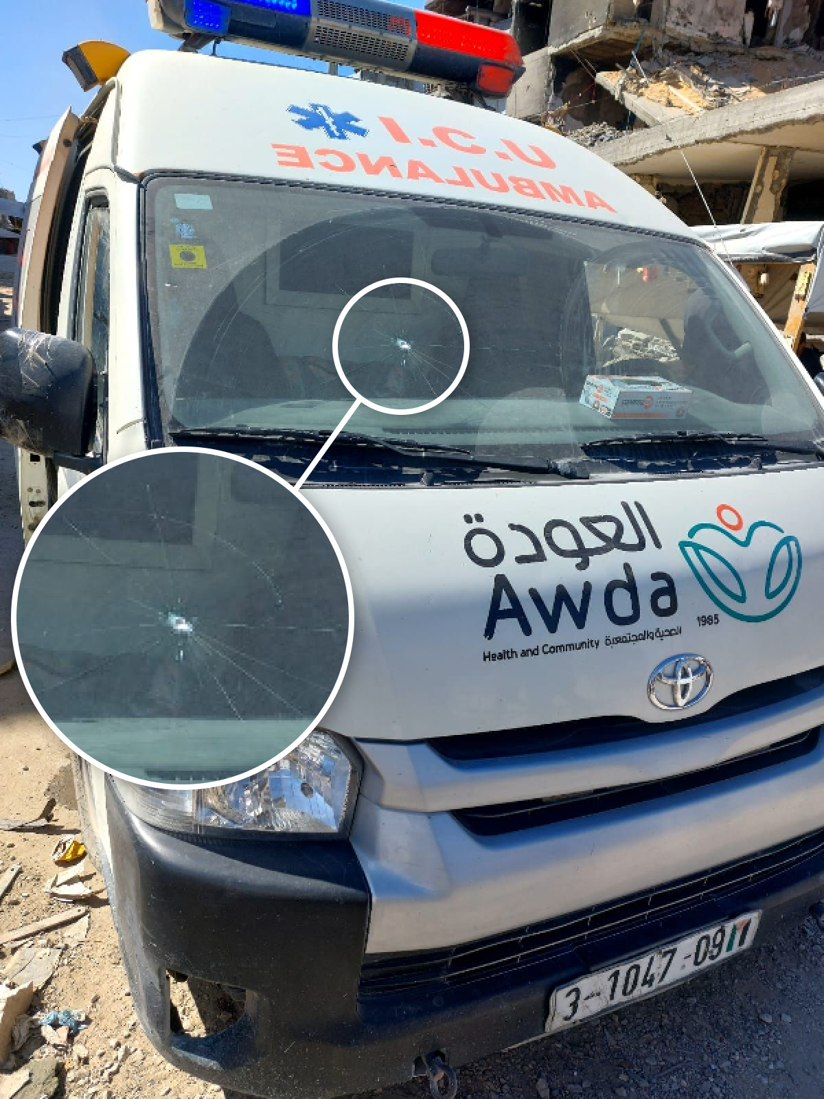 An ambulance is parked outside of a heavily damaged builting. The windscreen of the ambulance is damaged - we can see a small hole.