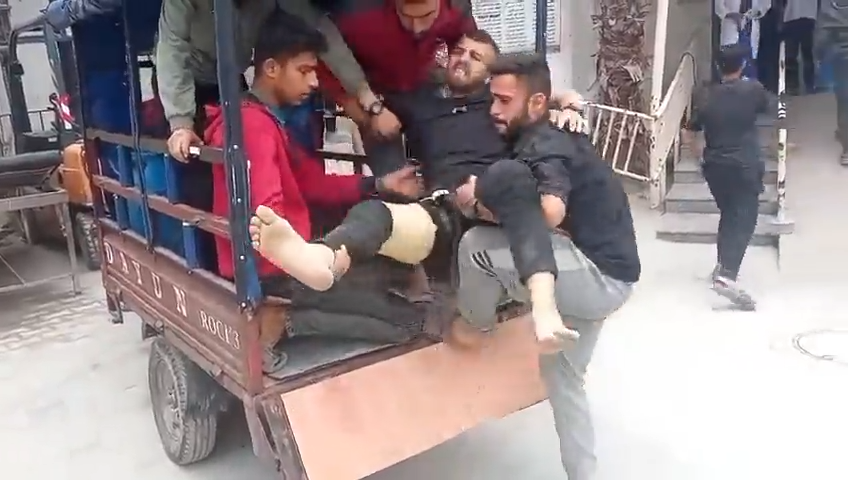 Several men help carry an injured man with a bandaged leg out of the back of a small vehicle, outside al-Awda hospital. The injured man appears to be in pain and is being supported by others as they assist him to the nearby building.