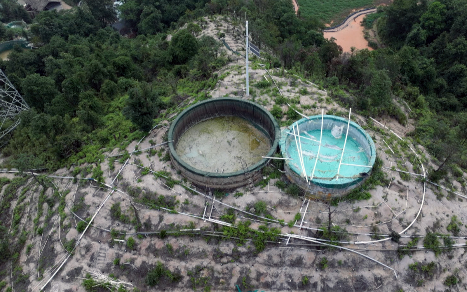 Vista aérea de dos grandes tanques de agua circulares en una ladera. Uno de ellos se ve fangoso y parcialmente lleno, mientras que el otro contiene agua azul claro con tuberías blancas rotas esparcidas por el suelo. La ladera es seca y desigual, con escasa vegetación, rodeada de un denso bosque verde.