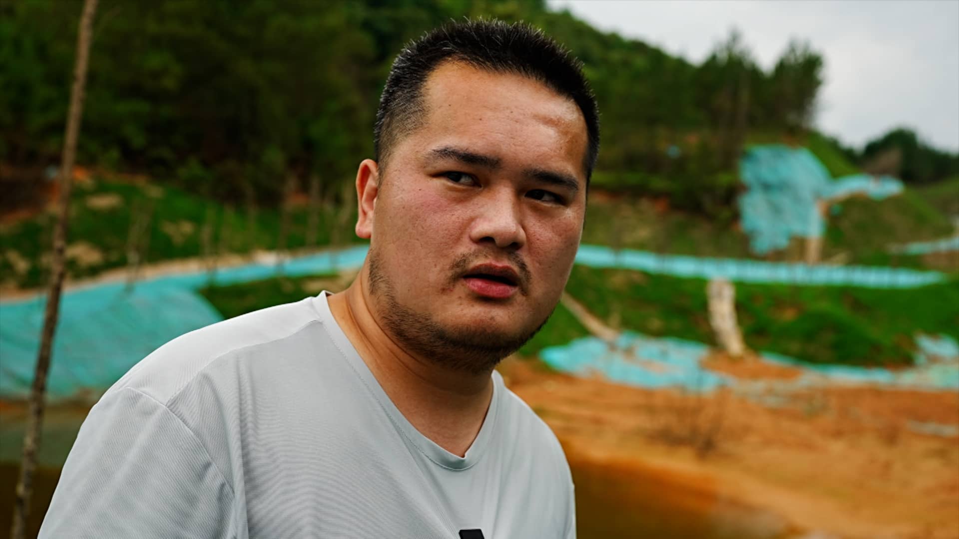 El agricultor chino Huang Xiaocong está junto a un depósito de arena con rastros de láminas de plástico turquesa y una exuberante vegetación en el fondo, y explica su difícil situación al vivir en una zona minera.