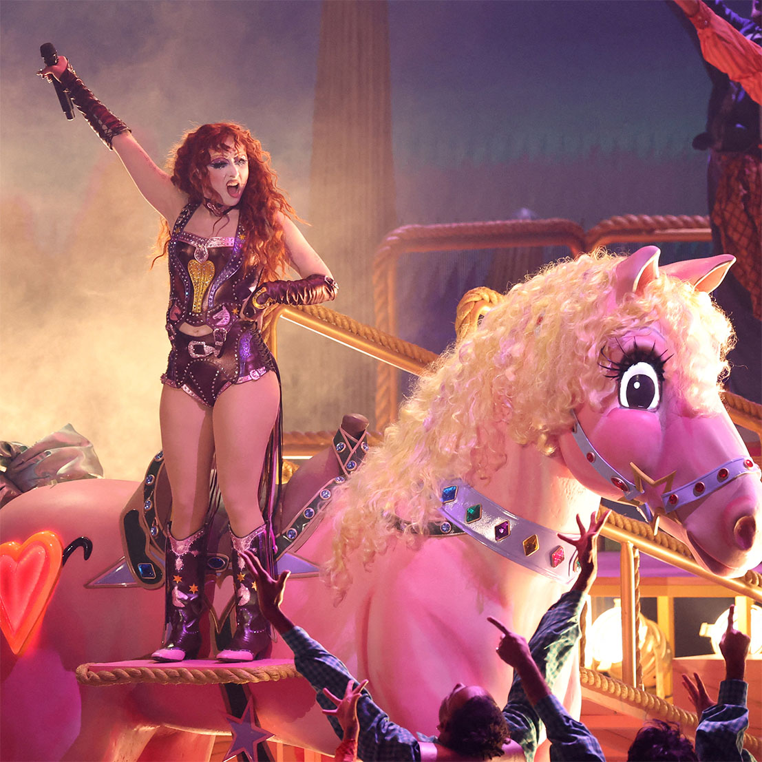 Chappell Roan on stage in a leather bustier, hot pants and cowboy boots, holding her microphone in the air while standing on the saddle of a giant pink pony.
