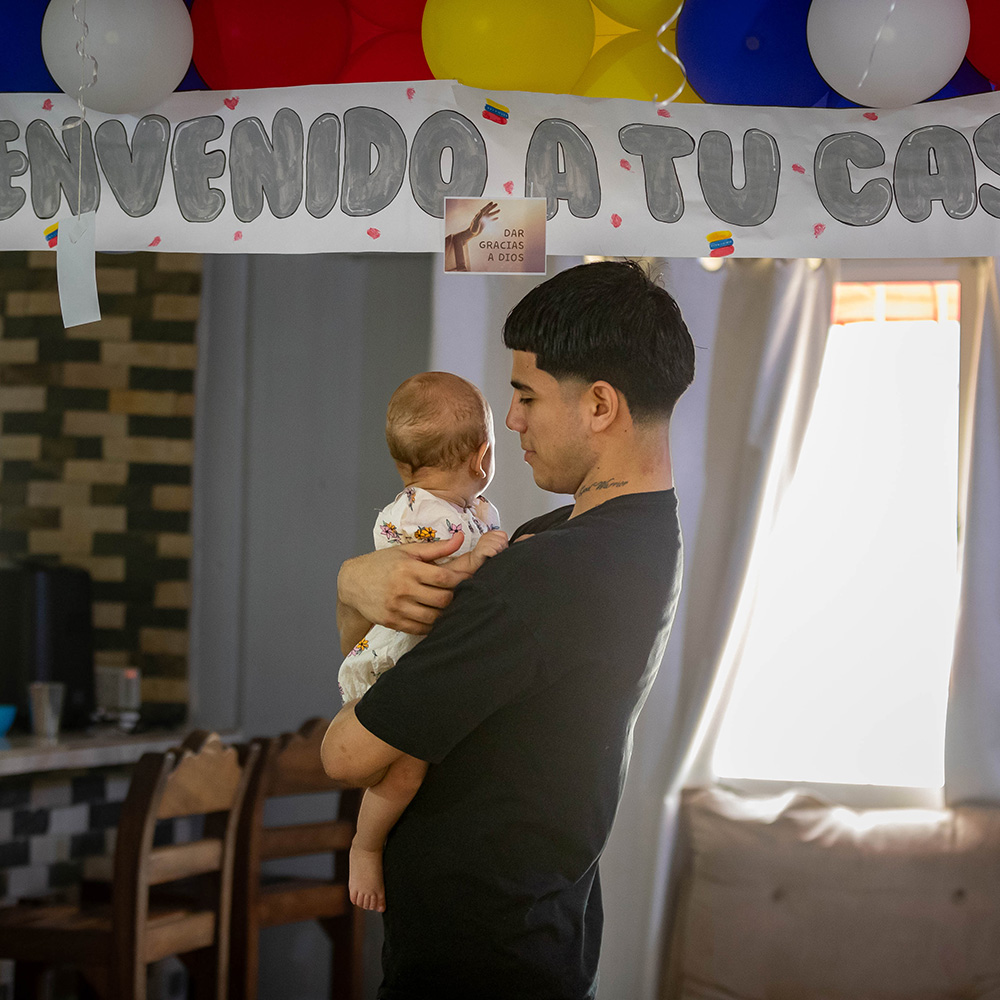 Joén Suarez, wearing a black shirt, holds his baby daughter in his house in Venezuela. In the background there are red, white, blue and yellow balloons and a sign saying "Welcome home" in Spanish