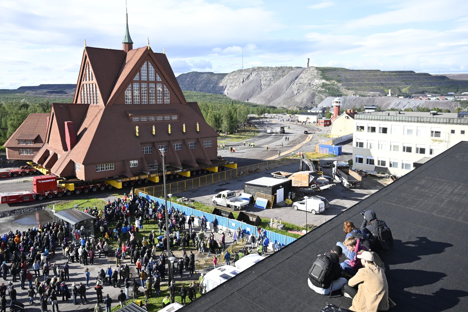 People gather to watch the moving of the wooden church, which can be seen on a platform with wheels on a rough road, with a grey stone landscape in the background and blue skies above