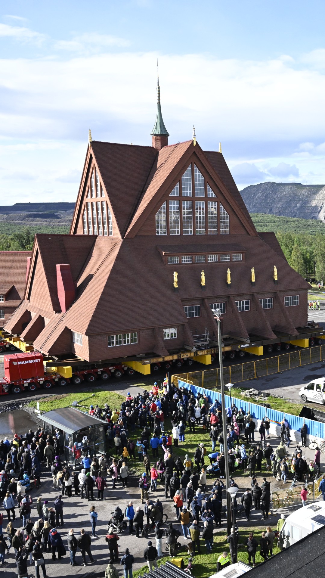 People gather to watch the moving of the wooden church, which can be seen on a platform with wheels on a rough road, with a grey stone landscape in the background and blue skies above