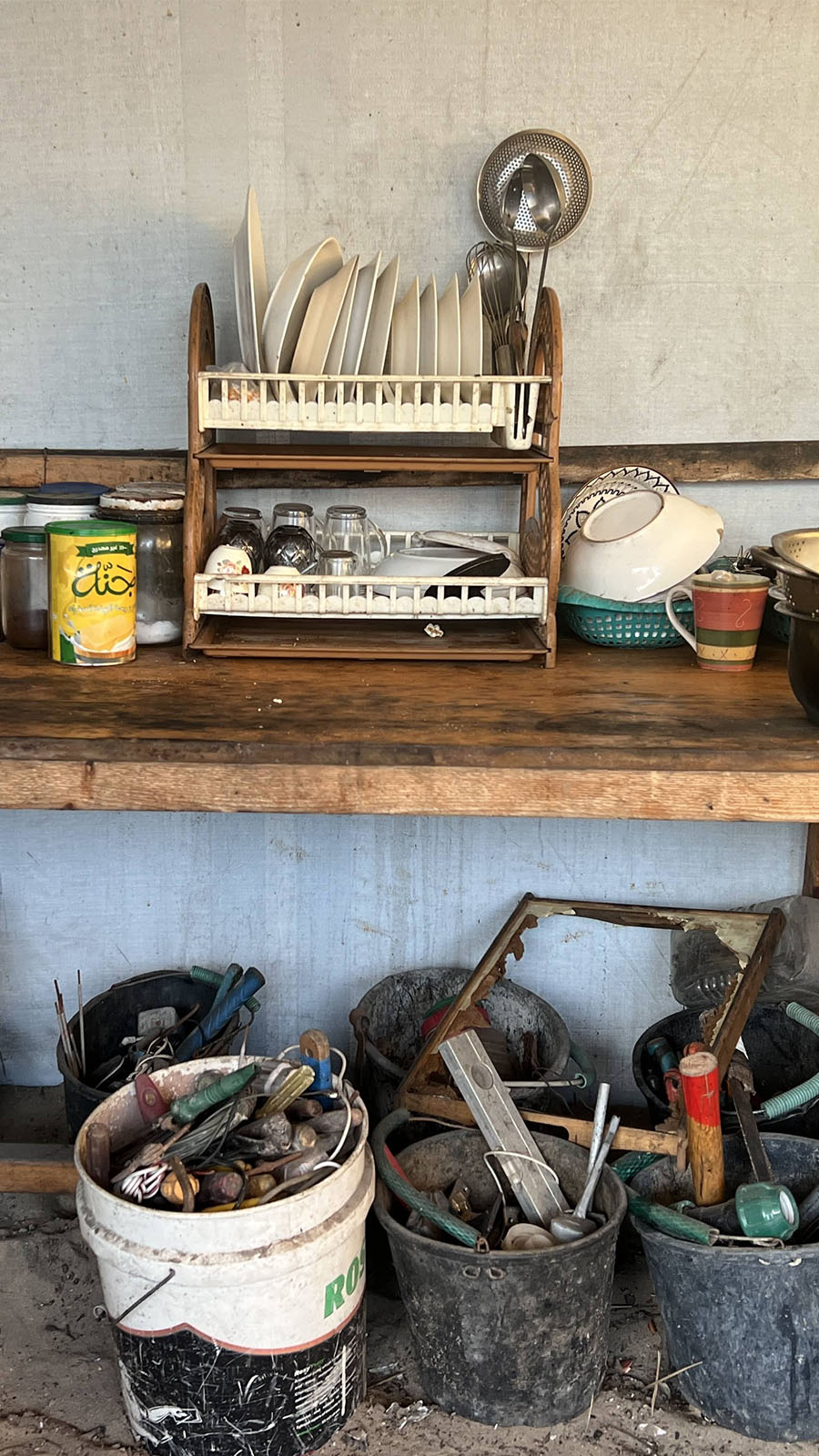 Mosab's rustic wooden kitchen table holding jars of spices, a bottle of oil, stacked dishes in a drying rack, cups, bowls, and metal pots. Below the table are plastic buckets filled with tools and household items