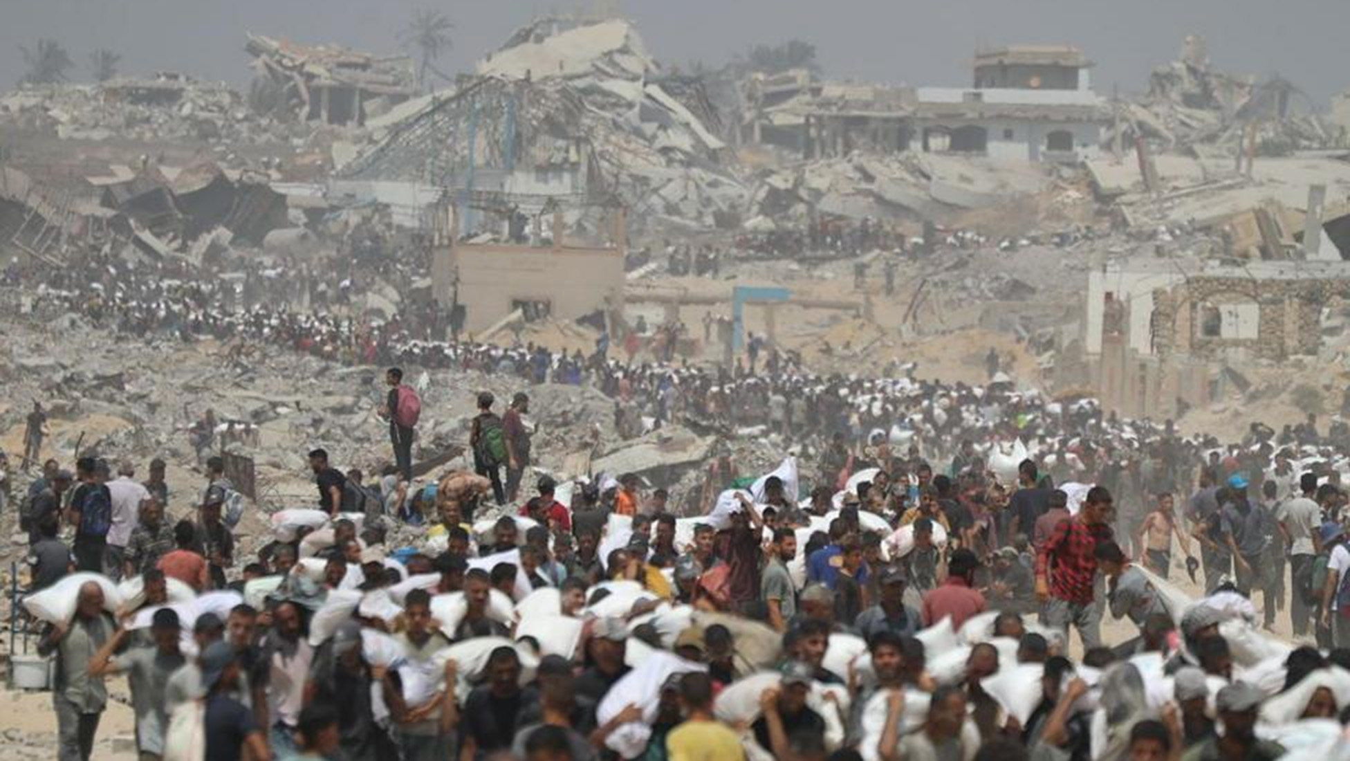 Geniş açıda önde Gazze'de çuvallarla yardım taşıyan insanlar görülüyor, arkada yıkılmış binalar var.