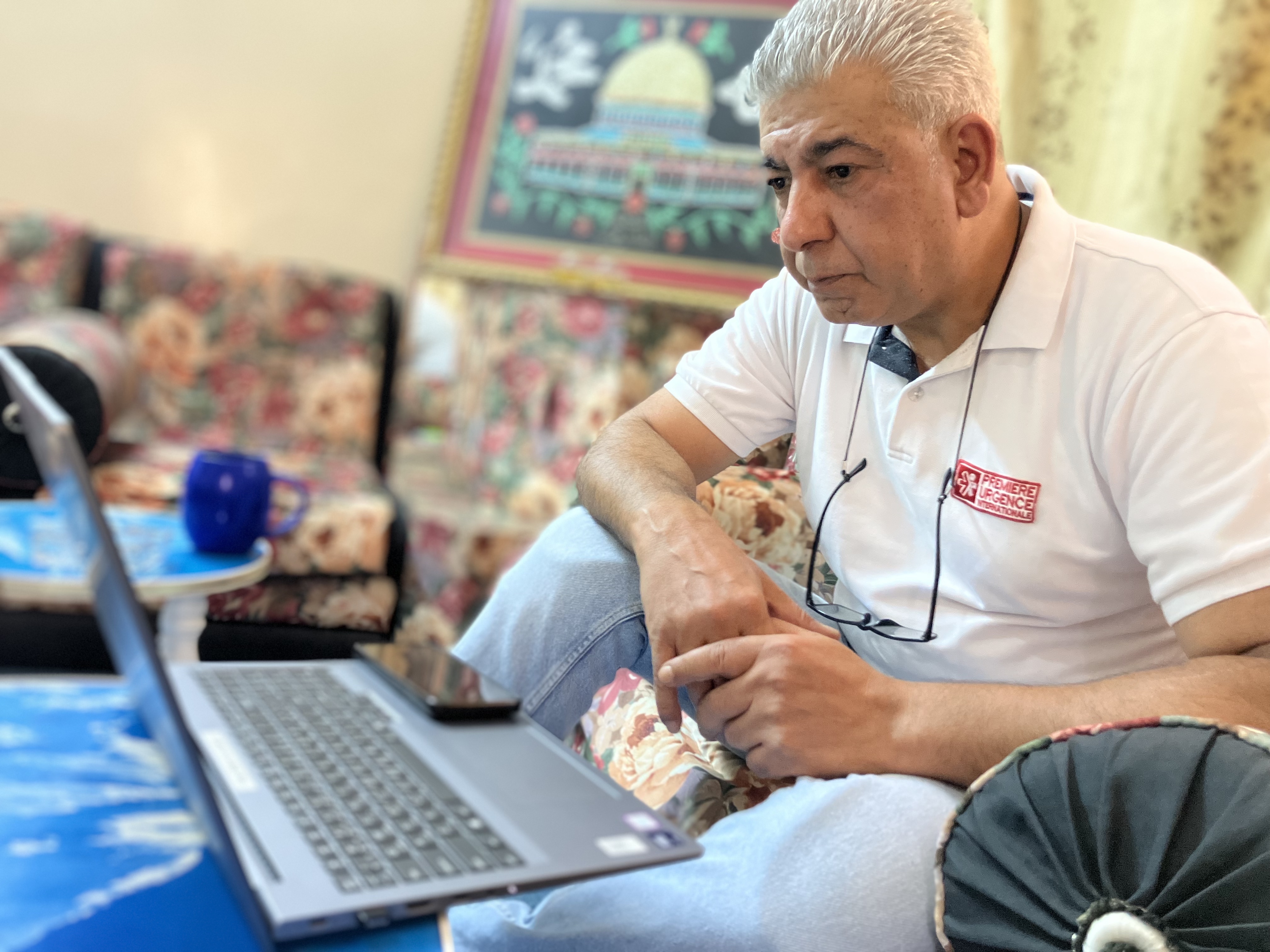 The photo shows Jehad Abu Hassan, wearing jeans and a white polo shirt, with glasses hanging from his neck, looking at a laptop while sitting on a floral sofa. In the background we can see two blue coffee tables, the floral seats and a framed painting of a mosque