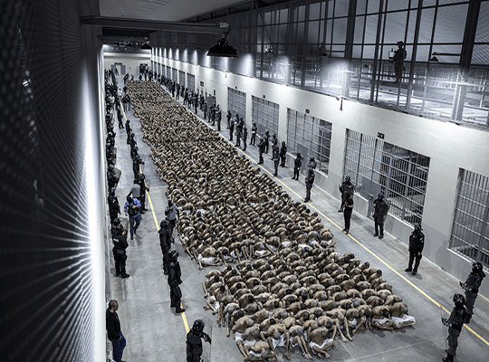 Prisioneros del Cecot sentados en fila rodeados de guardias de seguridad en uno de los pabellones de la cárcel