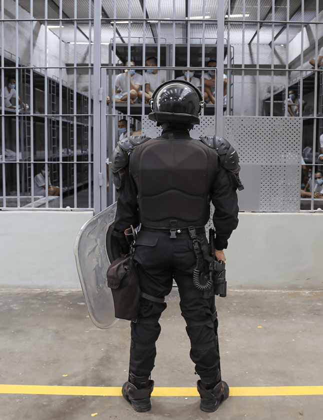 Un guardia del Cecot que se ve de espaldas, mira hacia una de las celdas en las que se ve a un grupo de reclusos