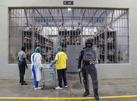 Dos guardias del Cecot junto a dos trabajadores del área de la salud frente a la entrada de una de las celdas donde se ve a un grupo de reclusos