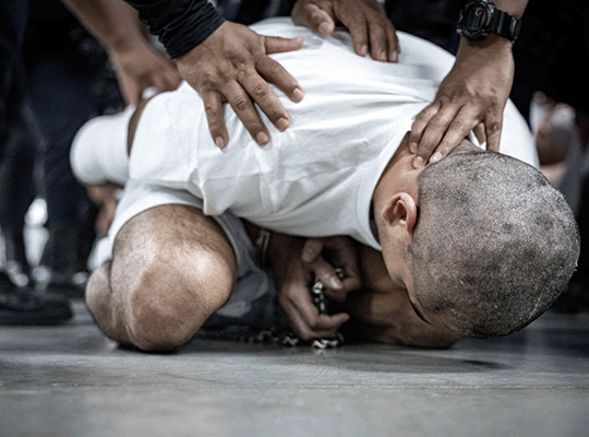 Un prisionero del Cecot arrodillado mientras varios guardias le ponen le ponen las manos sobre su espalda y cuello