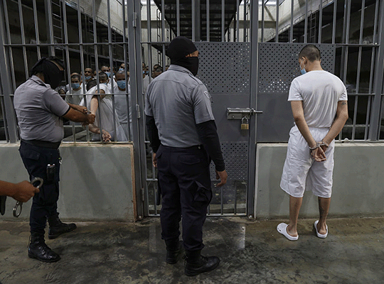 Dos guardias del Cecot con sus caras ocultas junto a un prisionero en la puerta de una celda y un grupo de prisioneros dentro de la celda