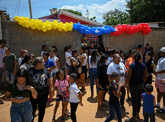 Un grupo de vecinos reunidos en una calle adornada con globos amarillos, azules y blancos para recibir a los migrantes venezolanos que estuvieron presos en el Cecot