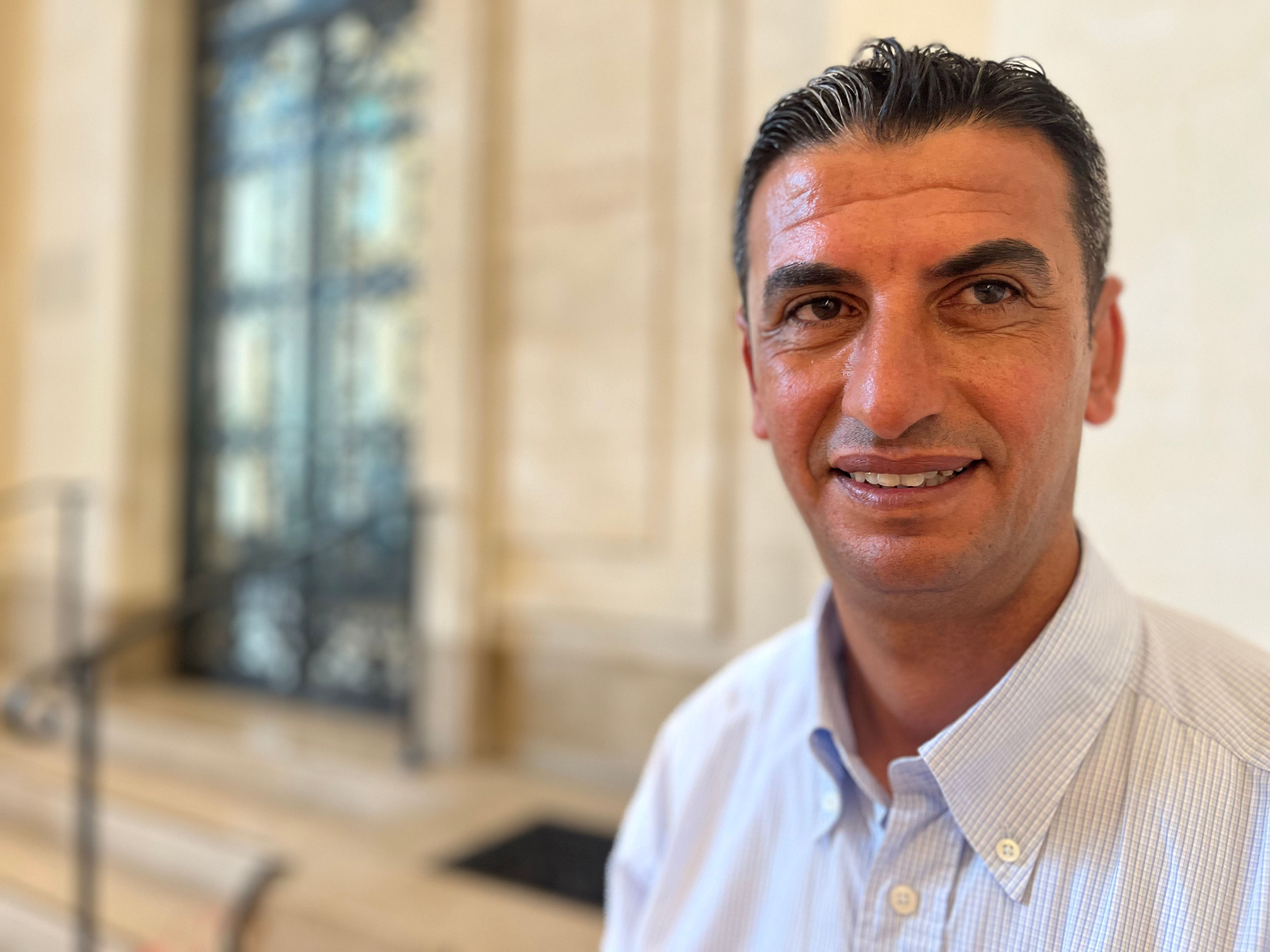 Archeologist Fadel al-Otol appears on the left corner of the image smiling and wearing a striped white and blue buttoned shirt. In the background, blurred, we can see the yellow walls of a building, stairs and a door