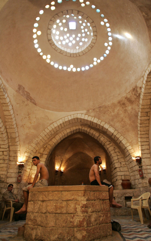 Two shirtless men sit on opposite sides of a stone structure in the Hammam a-Samra, inside the medieval gold market, in December 2005. In the background we can see the stone columns and arches of the building, lamps lighting the tunnels, ceramic vases on top of stone benches and a framed picture.