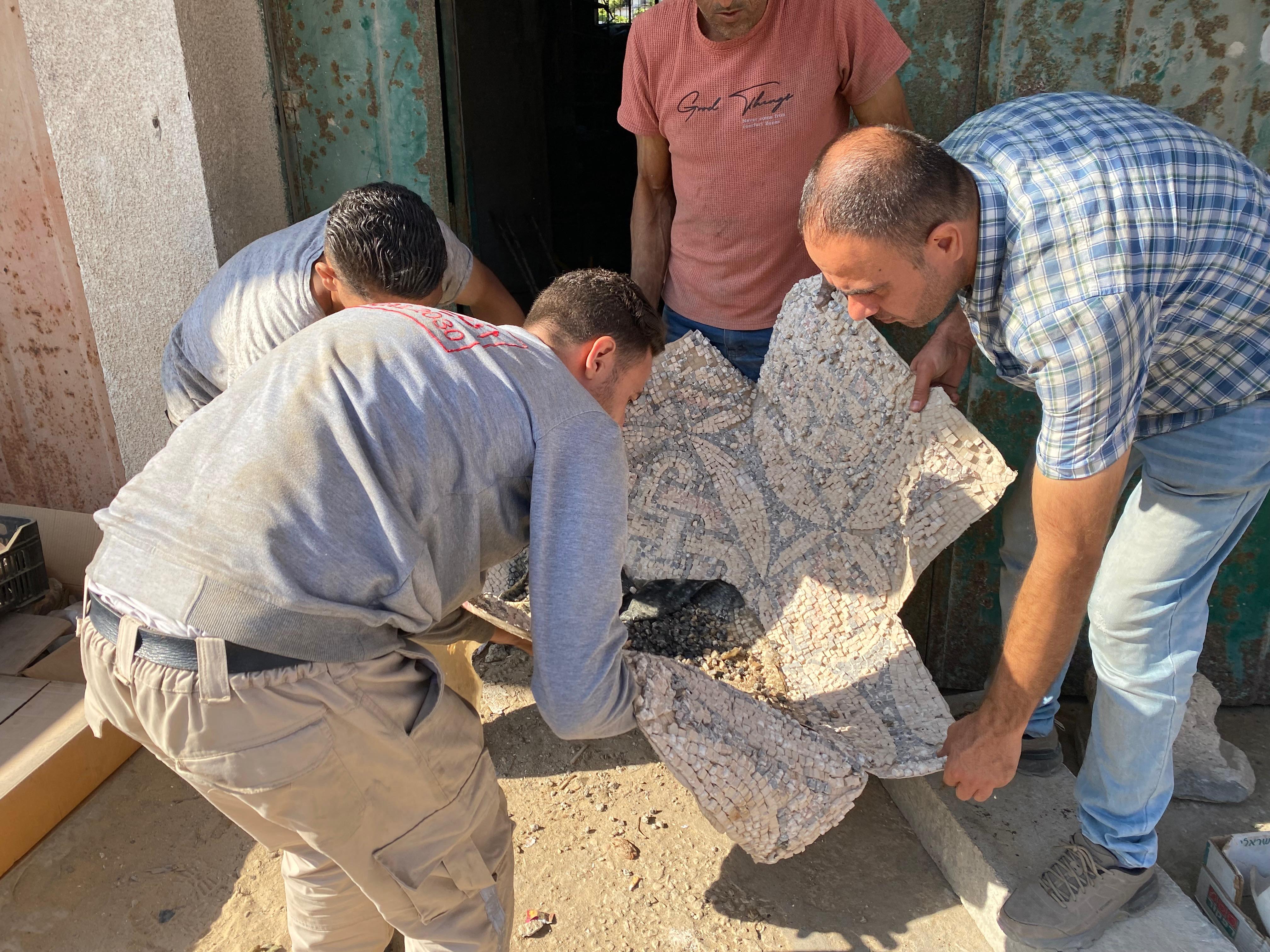 Image showing four volunteers carefully carry a colorful mosaic made of small tiles away from a building. The ground is dusty, and construction materials are scattered nearby