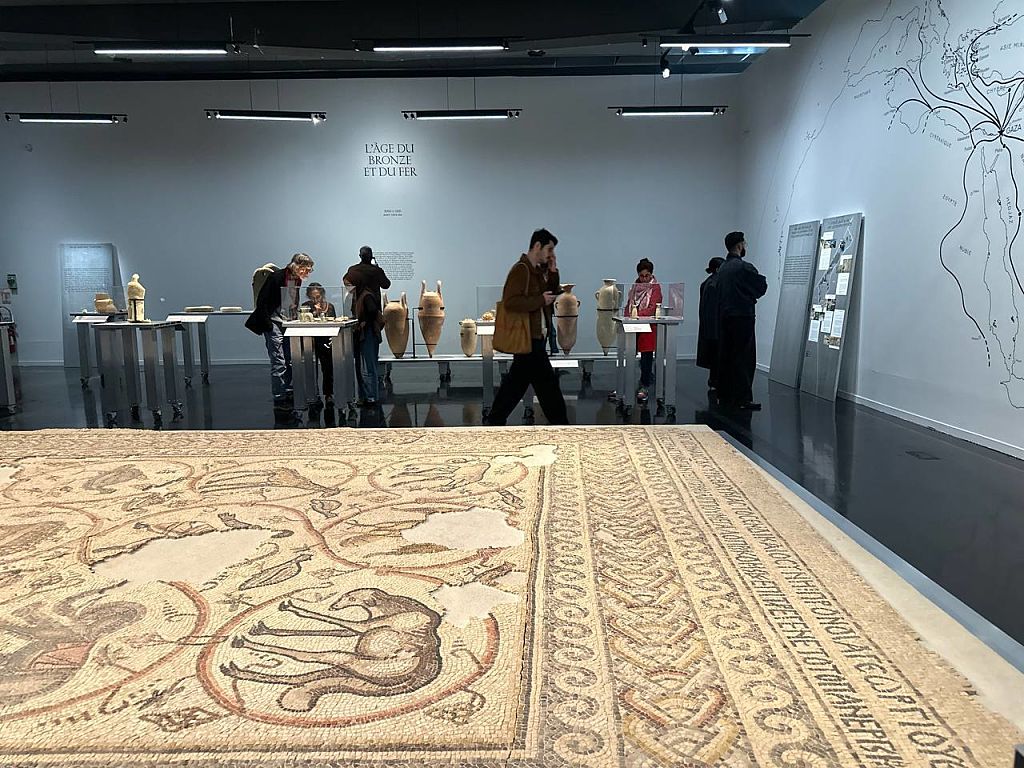 Museum exhibition room with visitors observing ancient artifacts. A large, detailed mosaic featuring animals and patterns covers the floor in the foreground. Display tables in the background hold pottery vases and other relics