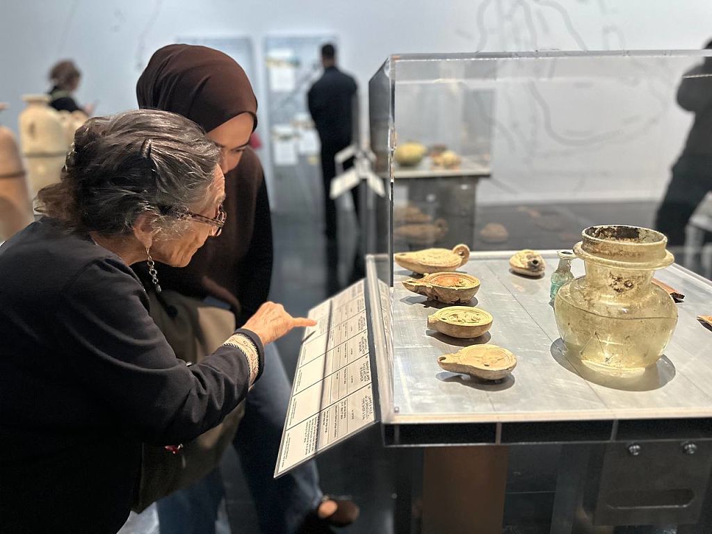 Two museum visitors closely examine a display case containing ancient artifacts, including oil lamps. One person points to an information panel beneath the case while the other observes attentively. Additional exhibits and visitors are visible in the background