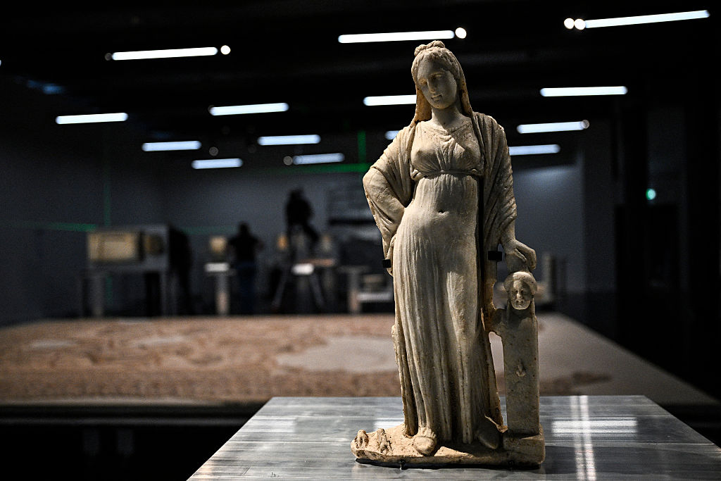 Marble statue of Aphrodite or Hecate, standing with one hand on her hip and the other resting on a small pillar. She is dressed in flowing draped garments that reach her feet. The statue is displayed indoors with dim lighting and other artifacts visible in the background