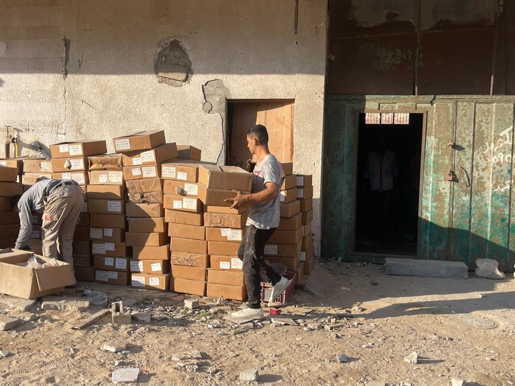 İki erkek tarihi eserlerle dolu kutuları hasarlı bir binanın dışına taşırken. Önlerinde yerde enkaz parçaları var.