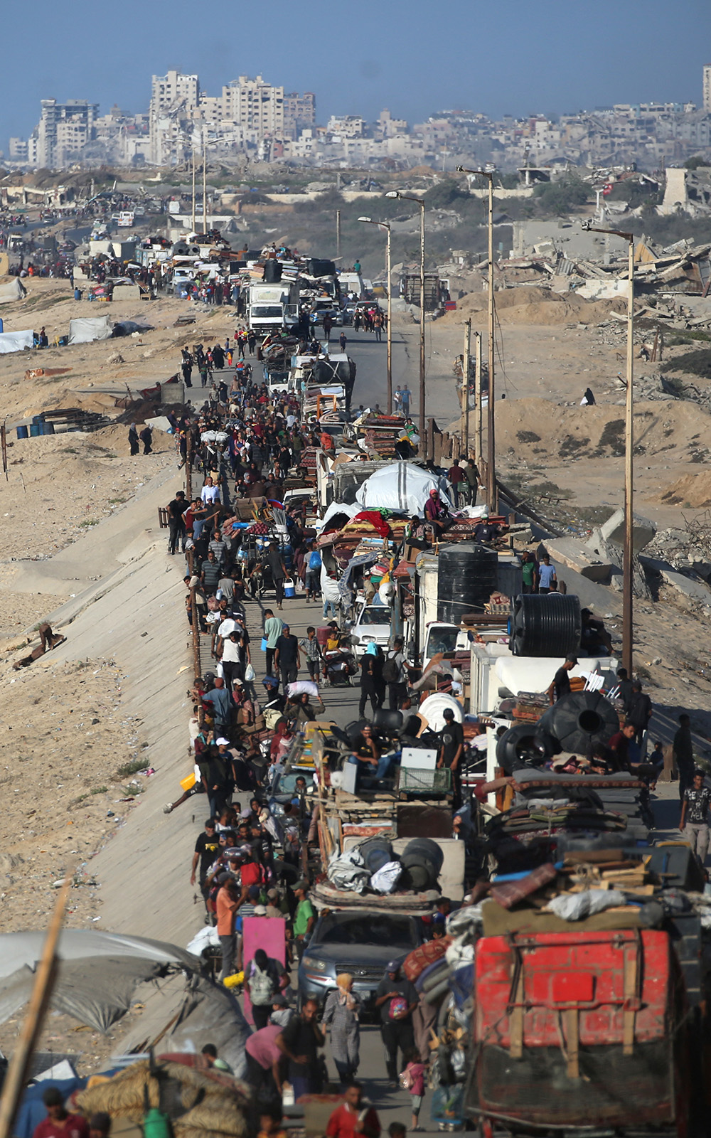 Displaced Palestinians move with their belongings southwards on a road in the Nuseirat refugee camp area in the central Gaza Strip following renewed Israeli evacuation orders for Gaza City on 16 September, 2025. Individuals walk alongside heavily loaded vehicles. The left side shows a beach, while the right side reveals damaged buildings and debris.