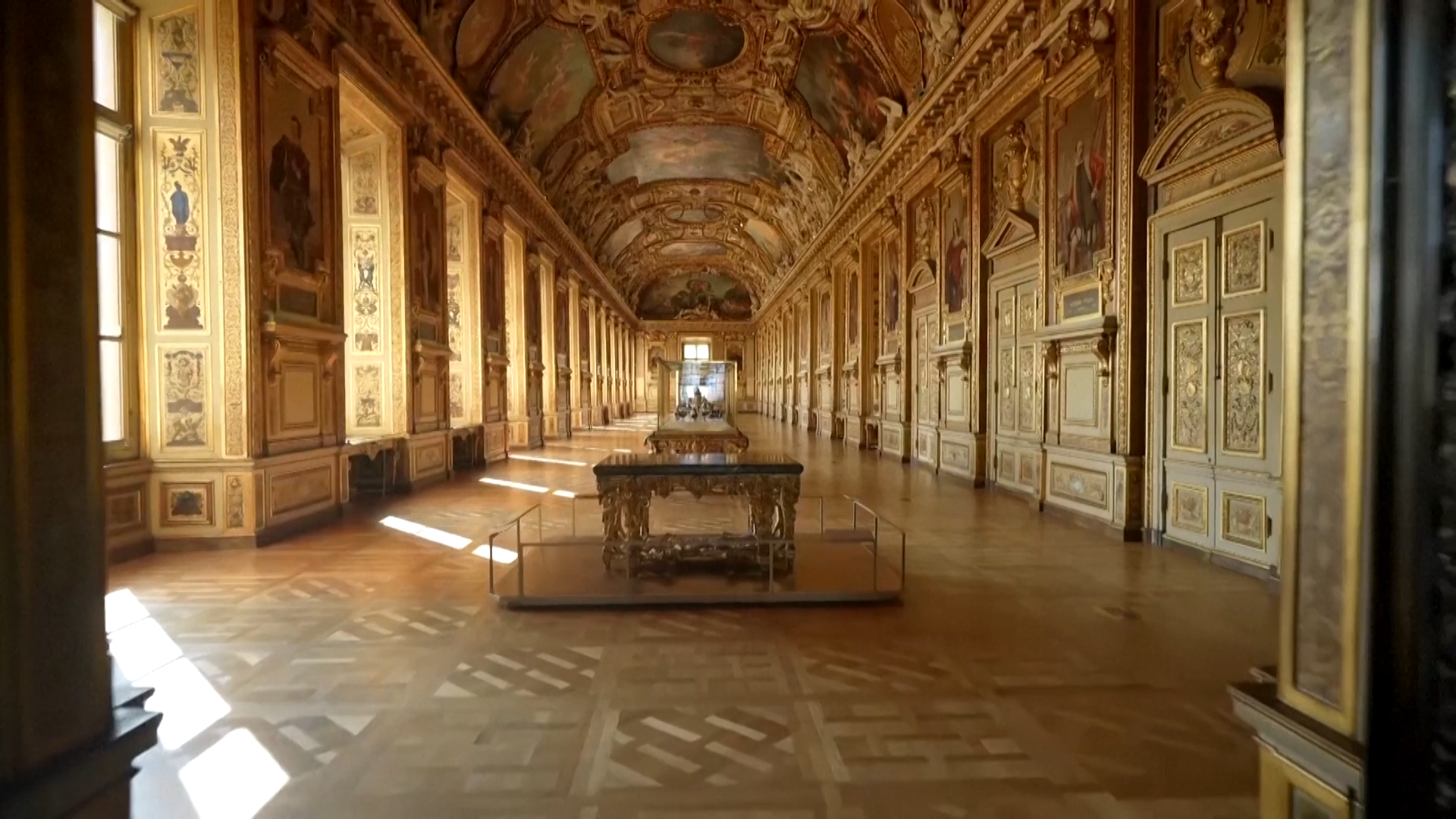 The interior of the Gallery of Apollo with tall windows along the left wall through which sunlight is falling onto the wooden parquet floor. Every surface is ornately decorated; the high-vaulted ceiling is painted with frescos and adorned with statues, and baroque gold leaf decorations flow down the walls, which also feature large portraits. In the centre of the room are a series of display cases. The three in the centre are tall and constructed from metal, other cases are more like tables with glass cases set on top. At the end of the room there is a large set of doors which are mostly glass, letting light flood into the room.