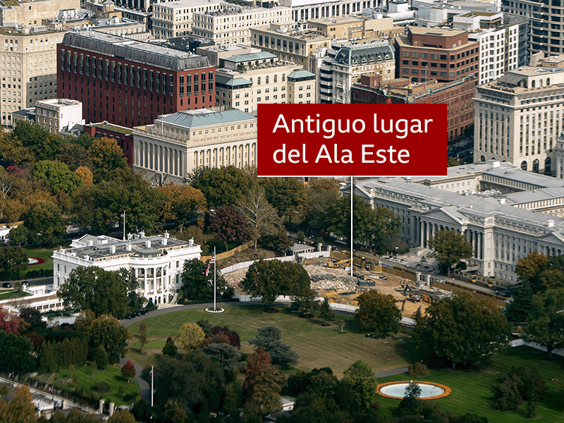 Vista aérea de los predios de la Casa Blanca que muestra el sitio de construcción despejado donde solía estar el Ala Este. El área está marcada con un rótulo rojo que lee “Antigua ubicación del Ala Este”. Alrededor del sitio hay árboles con follaje otoñal, edificios gubernamentales aledaños y la principal estructura de la Casa Blanca en primer plano.