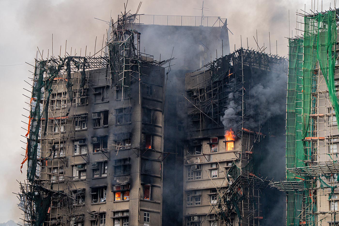 A photograph of one of the high-rise buildings in the Wang Fuk Court complex engulfed in flames and thick black smoke. The structure is covered in bamboo scaffolding, some of which is partially collapsed or charred. Bright orange flames are visible through several windows and along the exterior, while green safety netting hangs loosely on parts of the scaffolding. The sky is filled with dense smoke, obscuring the background.