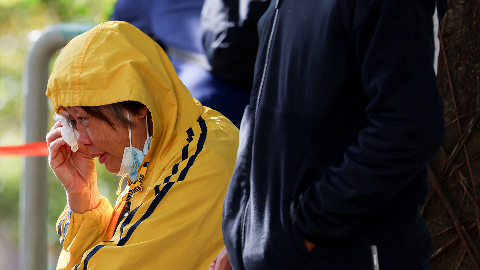 Woman dressed in a yellow hooded jacket wipes a tear from her eyes with a tissue as she sits next to others