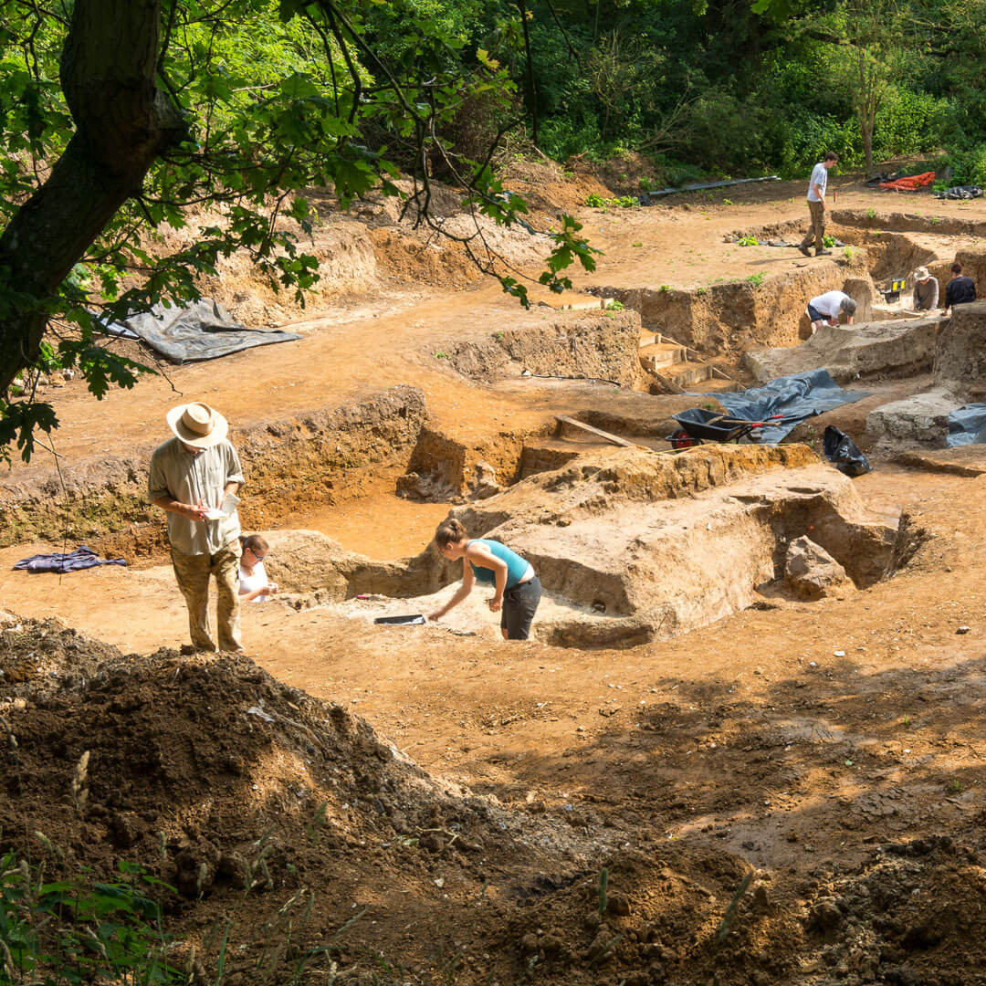 The area of the archaeological dig where the discovery was made, photographed on a sunny day through the shade of the surrounding trees. The site is in an open patch in a wooded area and made up of three or four connected pits in the yellow earth, each about two metres deep. Some of the site is covered in large black tarpaulin sheets. There are seven archaeologists standing in and around the pits. In the foreground is a man in a wide-brimmed brown hat and two women working in the nearest pit, one in a turquoise crop top and one in a white t-shirt and sunglasses.