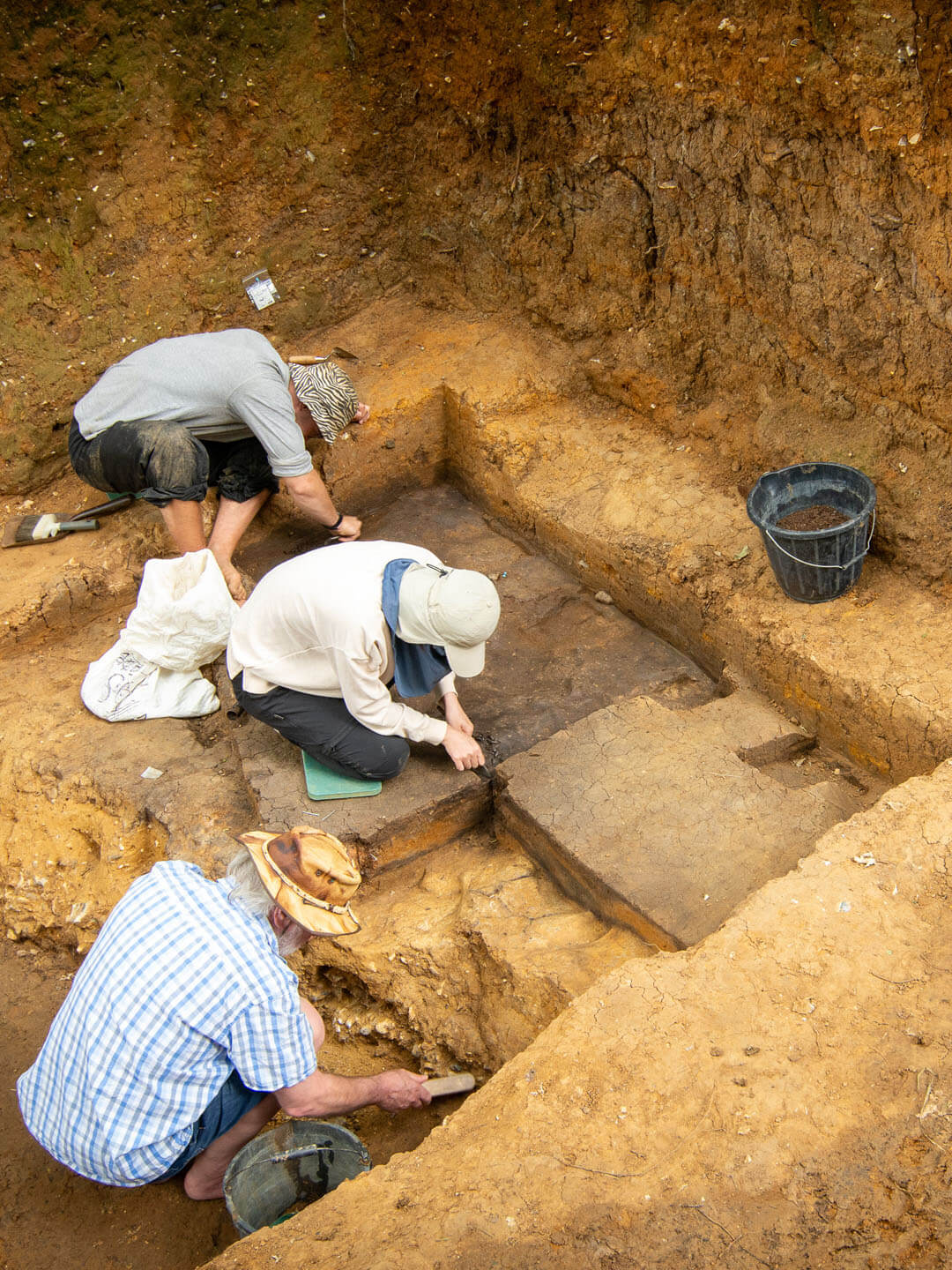 Three archaeologists working at the bottom of a pit with the walls looming above them. All three have their backs to us and are wearing hats. One has removed his sandals. All three are working on a square of earth, a little under two metres wide dug into the floor of the pit. They appear to be excavating a layer that is darker than the surrounding earth. 