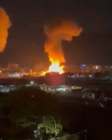 Photo of an explosion in a city the darkness with flame and smoke rising up. Some other buildings and trees are visible