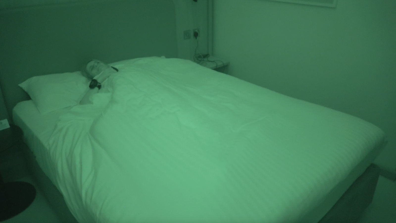 BBC reporter Ruth Clegg laying in bed asleep with sleeping equipment round her chest and nose. You can see her through an infrared camera showing it is night time.