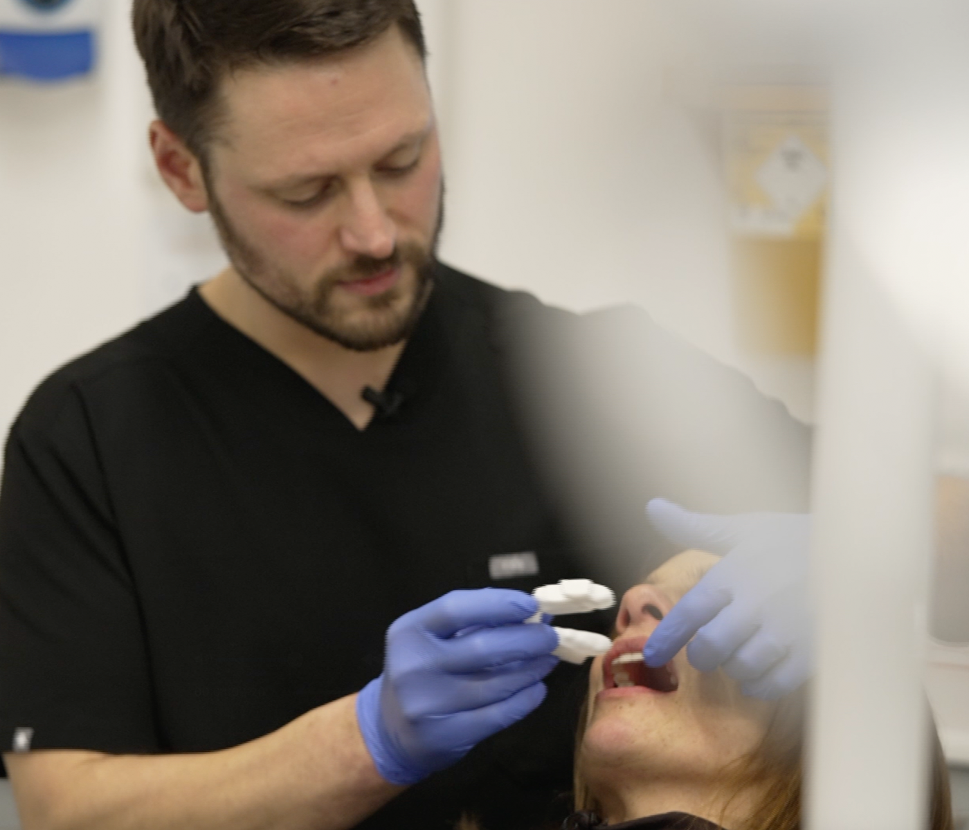 A dental professional wearing black scrubs and blue gloves places a mandibular advancement device into BBC reporter Ruth Clegg's mouth in a clinical setting.