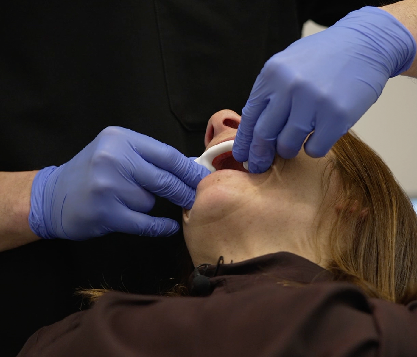 A close-up image of a dental professional wearing black scrubs and blue gloves places a mandibular advancement device into BBC reporter Ruth Clegg's mouth in a clinical setting.