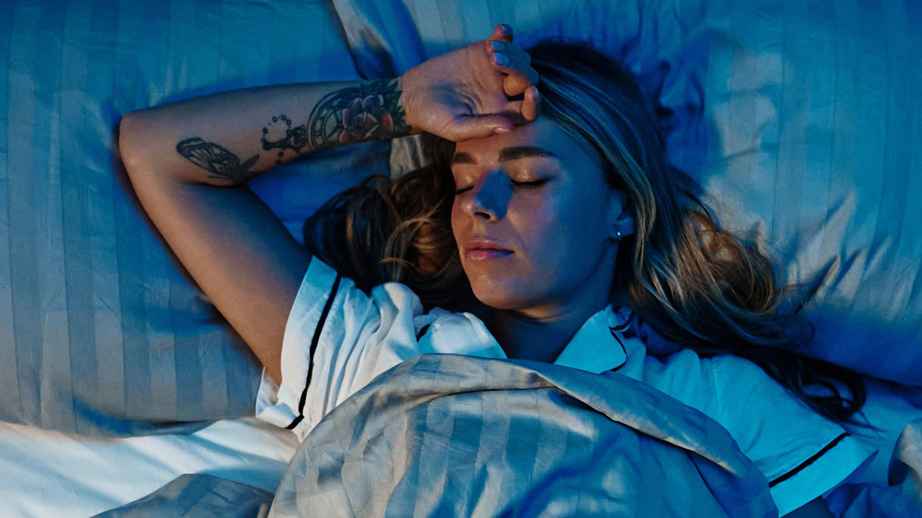 A young woman lying in bed trying to get to sleep, she has her right hand against her forehead revealing a snaking tatoo on her wrist.
