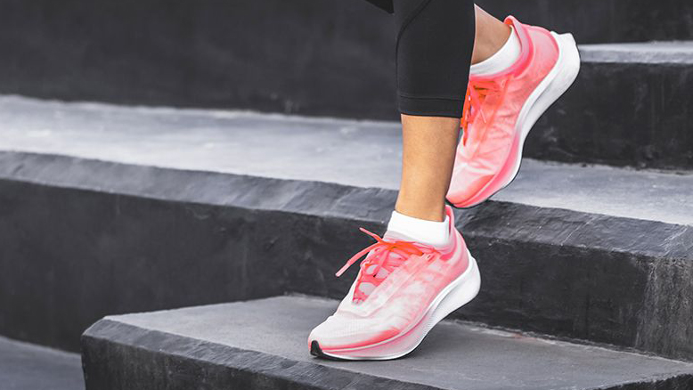 A woman wearing black leggings and pink athletic shoes, standing on a set of black steps. One foot is placed on a higher step while the other remains on a lower one, suggesting an exercise or stretching.