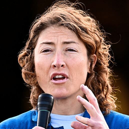 A headshot of Christina Koch talking into a microphone. Her left hand is raised in front of her and the wind is blowing her hair as she talks.