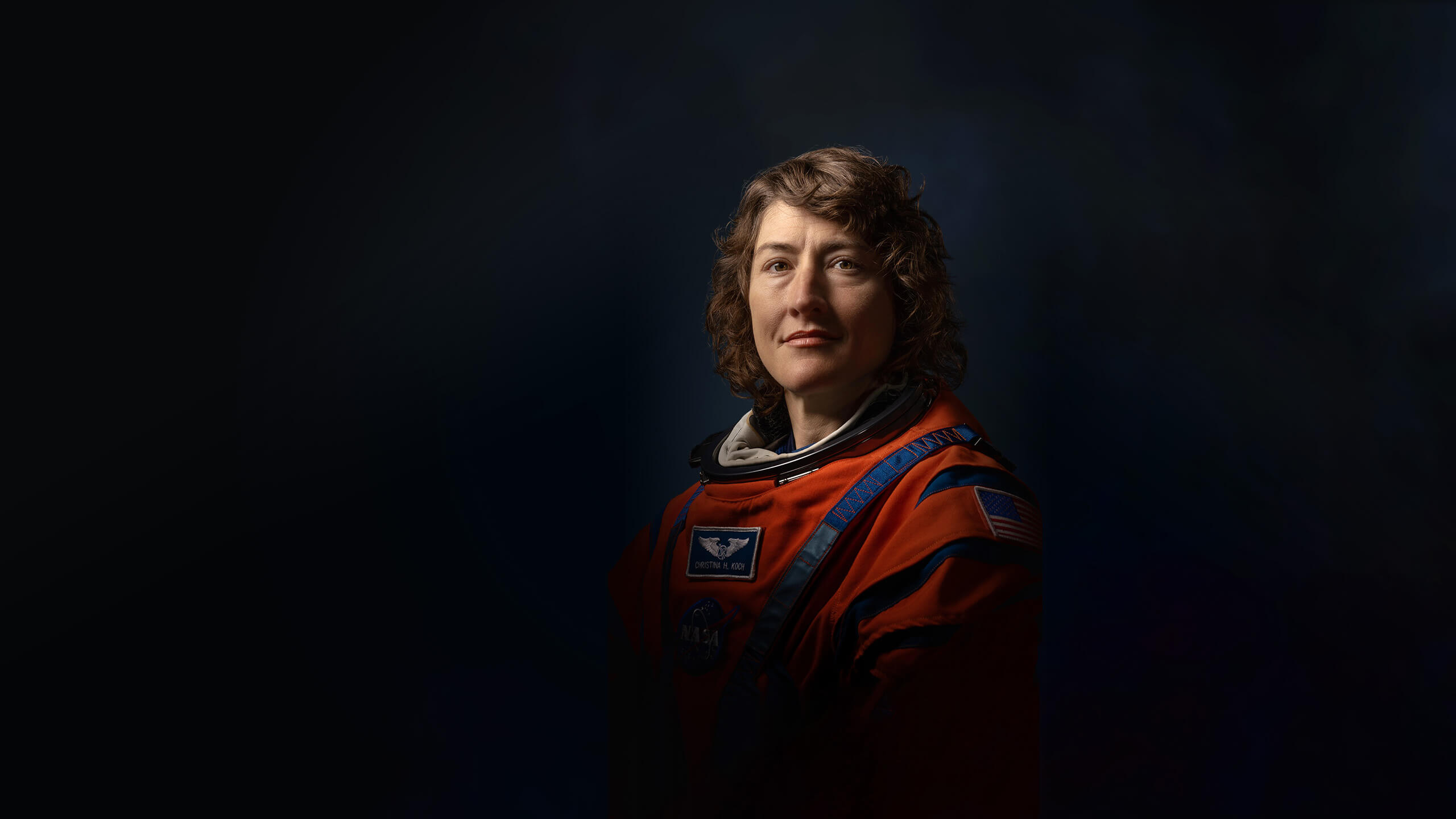 Official Nasa portrait of Christina Koch in her orange spacesuit showing her from mid-waist. The lighting is high contrast and dramatic making him look heroic. Her hair falls in curls either side of her face. She stares towars the camera looking both contemplative and determined.