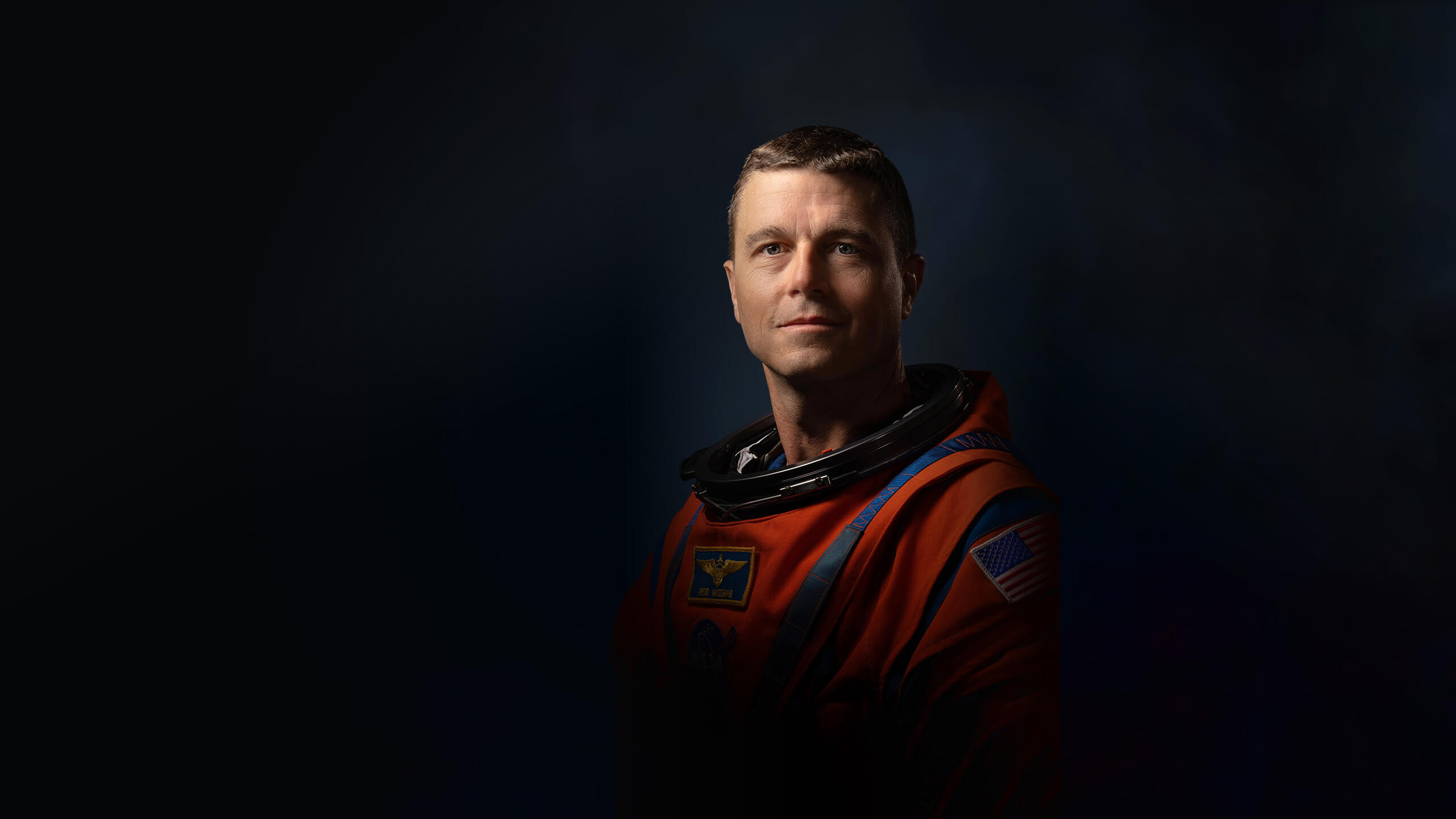 Official Nasa portrait of Reid Wiseman in his orange spacesuit showing him from mid-waist. The lighting is dramatic with the right side of his face in bright light and shadows cast across the other side making him look heroic but there is a slight smile threatening to break across his face.