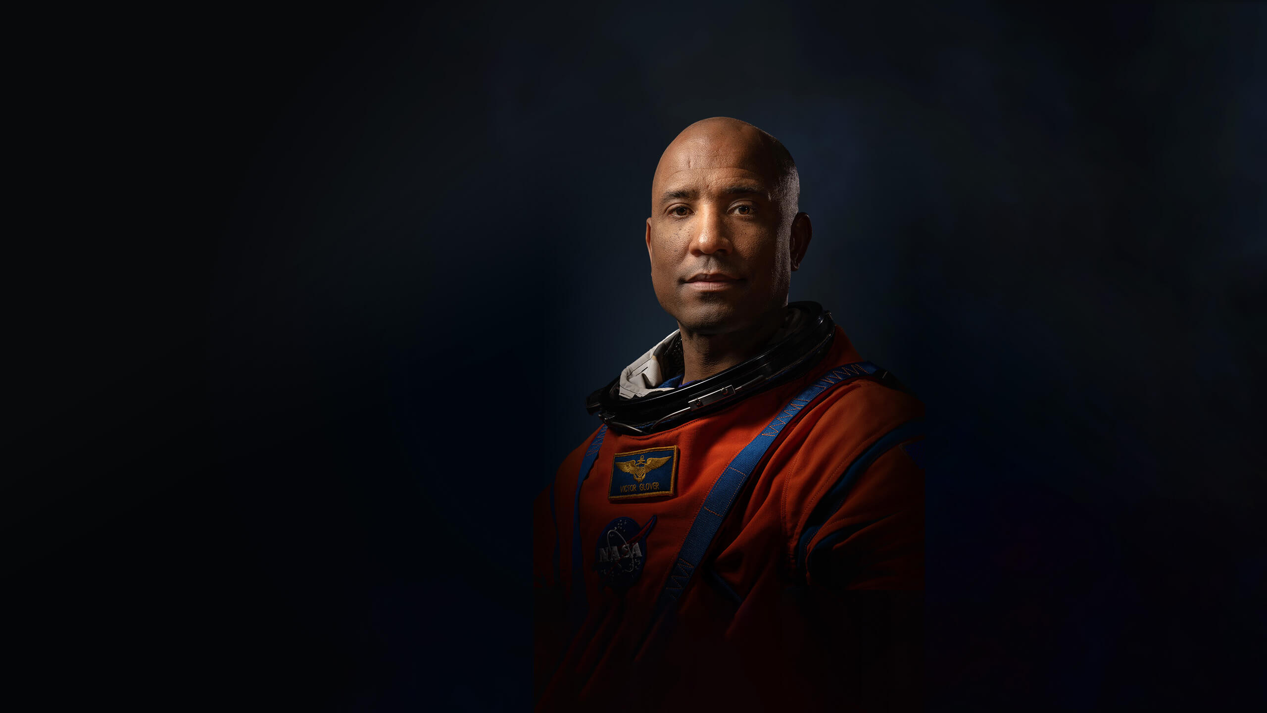 Official Nasa portrait of Victor Glover in his orange spacesuit showing him from mid-waist. The lighting is high contrast and dramatic making him look heroic. He is looking directly at the camera with a stoic expression.
