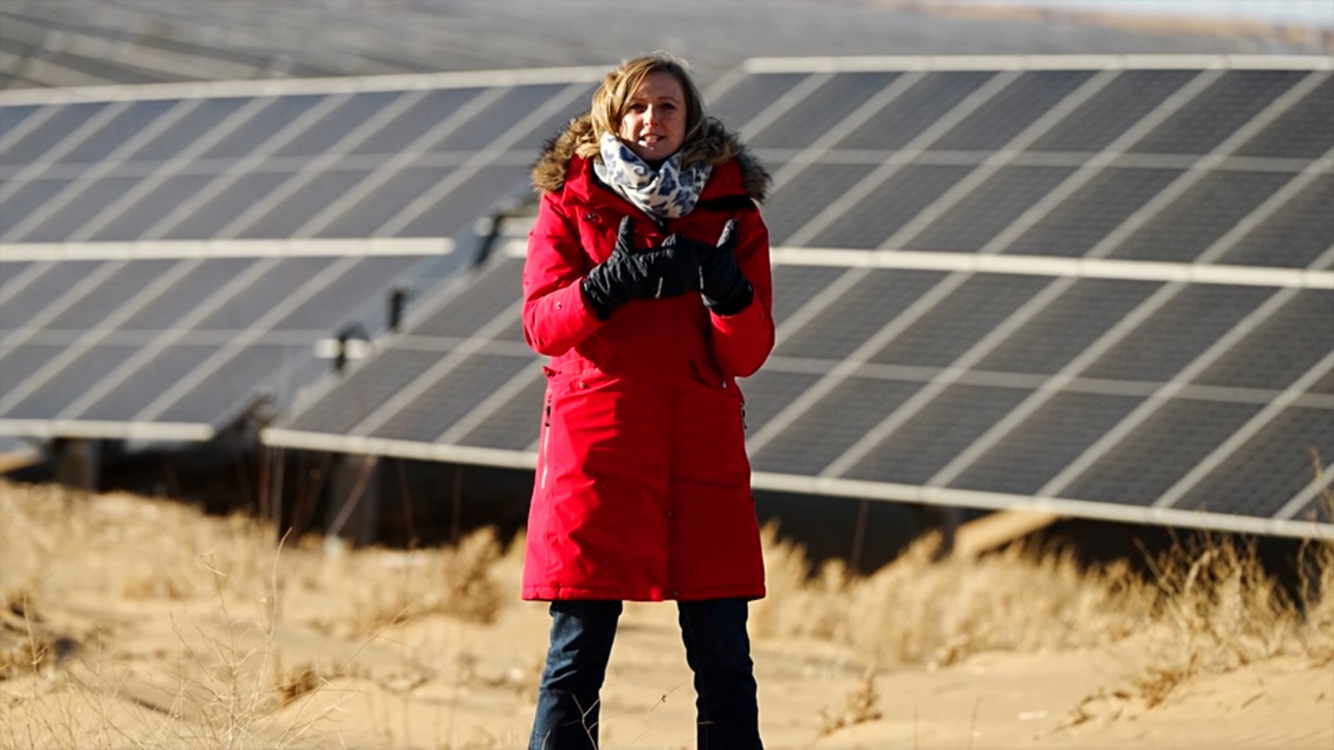 碧嘉蘭身穿厚重紅色外套、深色長褲與手套，站在內蒙古沙漠大片太陽能板前