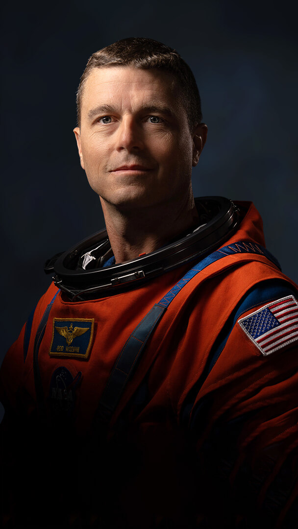 Official Nasa portrait of Reid Wiseman in his orange spacesuit showing him from mid-waist. The chiaroscuro lighting from a high angle illuminates the right side of his face and casts shadows across the left side. There is a slight smile threatening to break across his face.